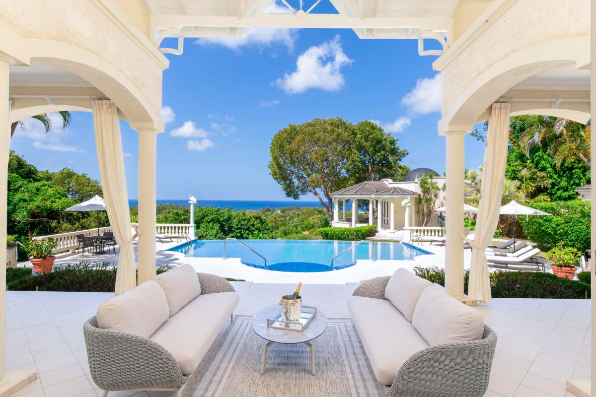 A luxurious outdoor patio at Sandy Lane luxury villa features two white sofas and a glass table overlooking an infinity pool, lush greenery, and the ocean under a bright blue sky with fluffy clouds.