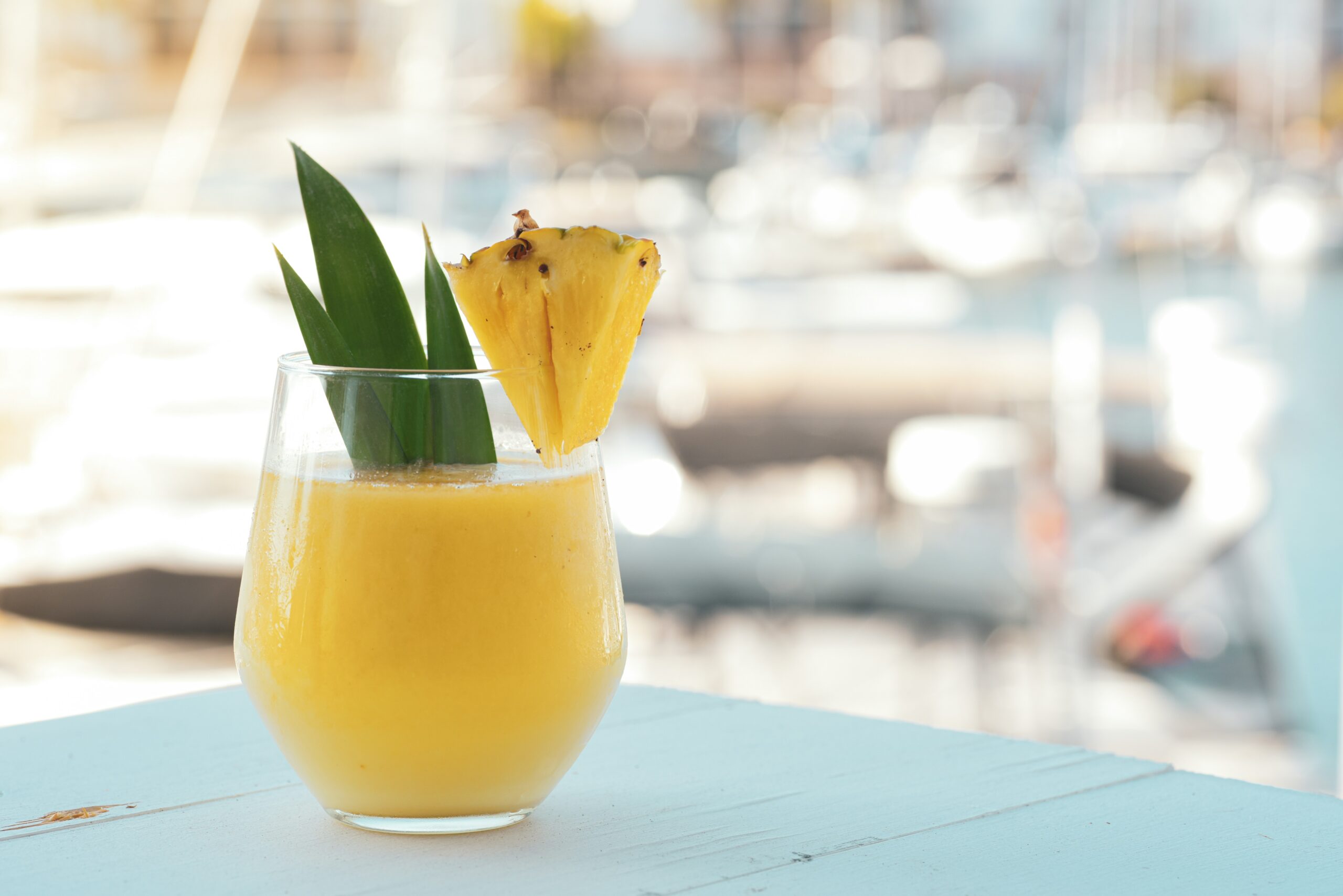 Pictured is a blended Pina Colada, one of the best Caribbean cocktails and most widely-known, sitting on a table by the beach with a chunk of pineapple on the rim and a pineapple frond used as garnish on the drink.