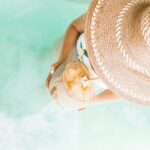 Woman drinks one of the best Caribbean drinks out of a coconut shell on her vacation on the Caribbean, walking into the clear blue water with a hat on her head.