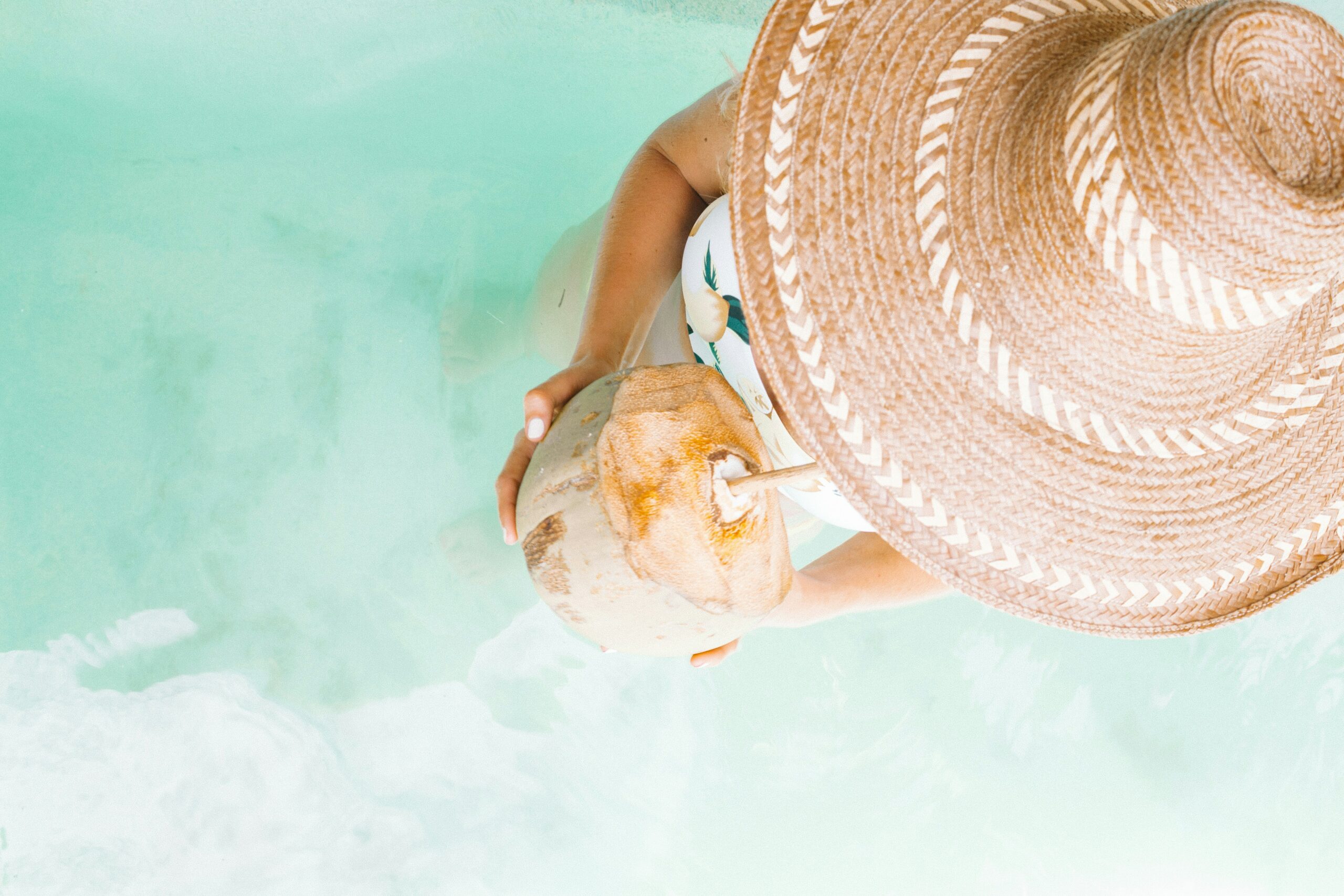 Woman drinks one of the best Caribbean drinks out of a coconut shell on her vacation on the Caribbean, walking into the clear blue water with a hat on her head.