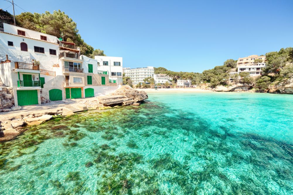 A picturesque coastal scene showcases one of the top 5 beaches in Mallorca, with clear turquoise water and rocky shores. Colorful buildings with green doors and shutters line the coast, backed by lush trees. In the distance, more charming architecture and a sandy beach are visible under a blue sky.