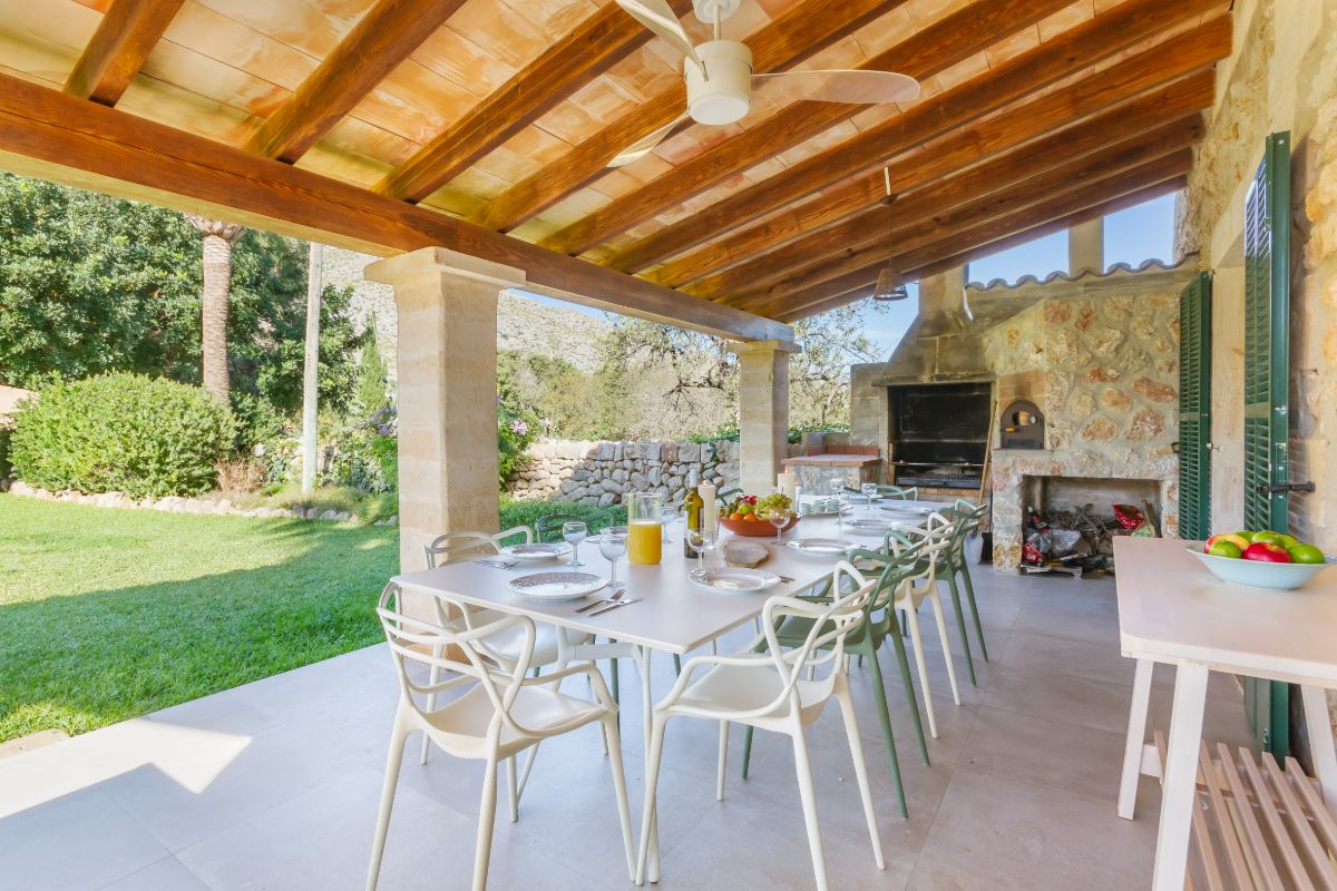 A covered patio at Can Fontana showcases a wooden ceiling and stone pillars, with a long dining table set with plates, glasses, and a pitcher of orange juice. In the background, a built-in outdoor grill enhances the inviting space, surrounded by a grassy yard and trees.