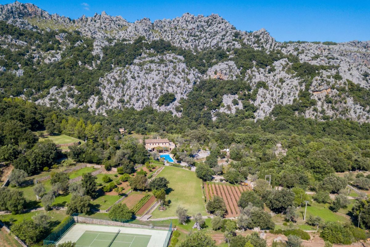 Aerial view of Can Sa Calma, a rural estate with gardens, a tennis court, a swimming pool, and a farmhouse, surrounded by dense greenery and rugged mountains under a clear blue sky.