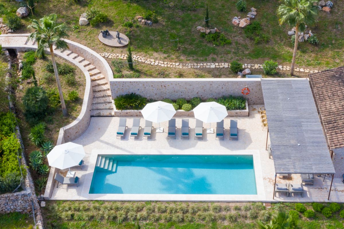 Aerial view of Can Rocaterra’s rectangular swimming pool with steps, surrounded by lounge chairs and white umbrellas. A curved stone staircase, lush greenery, and a shaded patio area complete this inviting oasis.