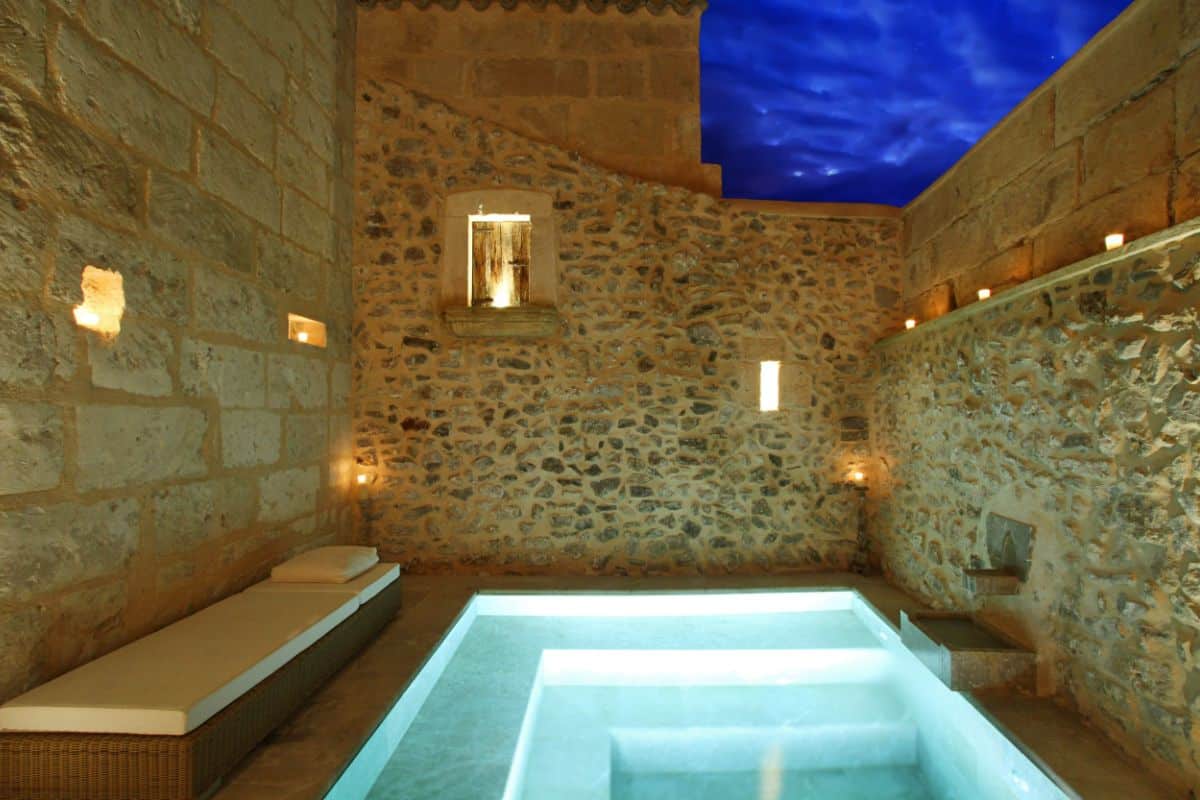 A small, illuminated indoor pool with stone walls, candles, and a cushioned bench creates a cozy, tranquil spa atmosphere—like an Estrella Mallorca retreat—under a deep blue evening sky.
