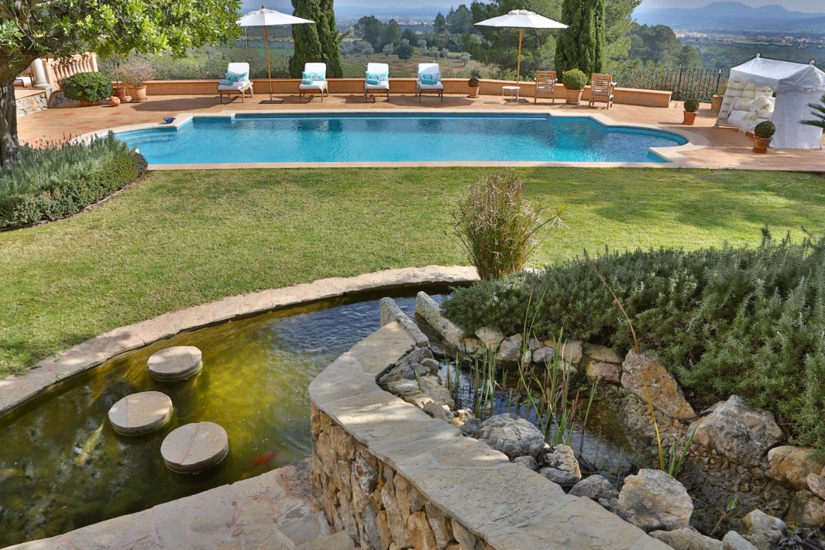 A landscaped backyard at Residencia Llum features a swimming pool, lounge chairs, umbrellas, and stunning mountain views. In the foreground, a small pond with stepping stones and rocks sits beside a stone staircase.