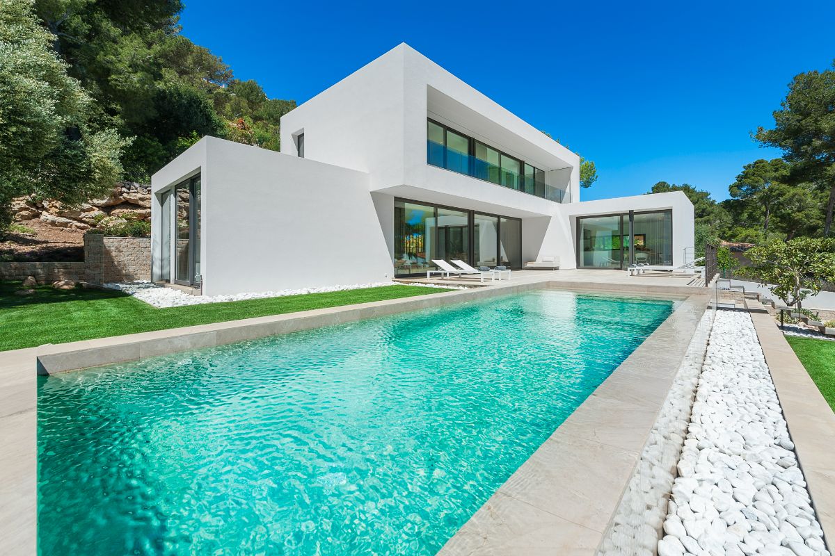 Modern white Villa Cala Brava with large glass windows, surrounded by green lawns and trees, featuring a rectangular swimming pool with clear turquoise water, sun loungers, and a stone border under a bright blue sky.