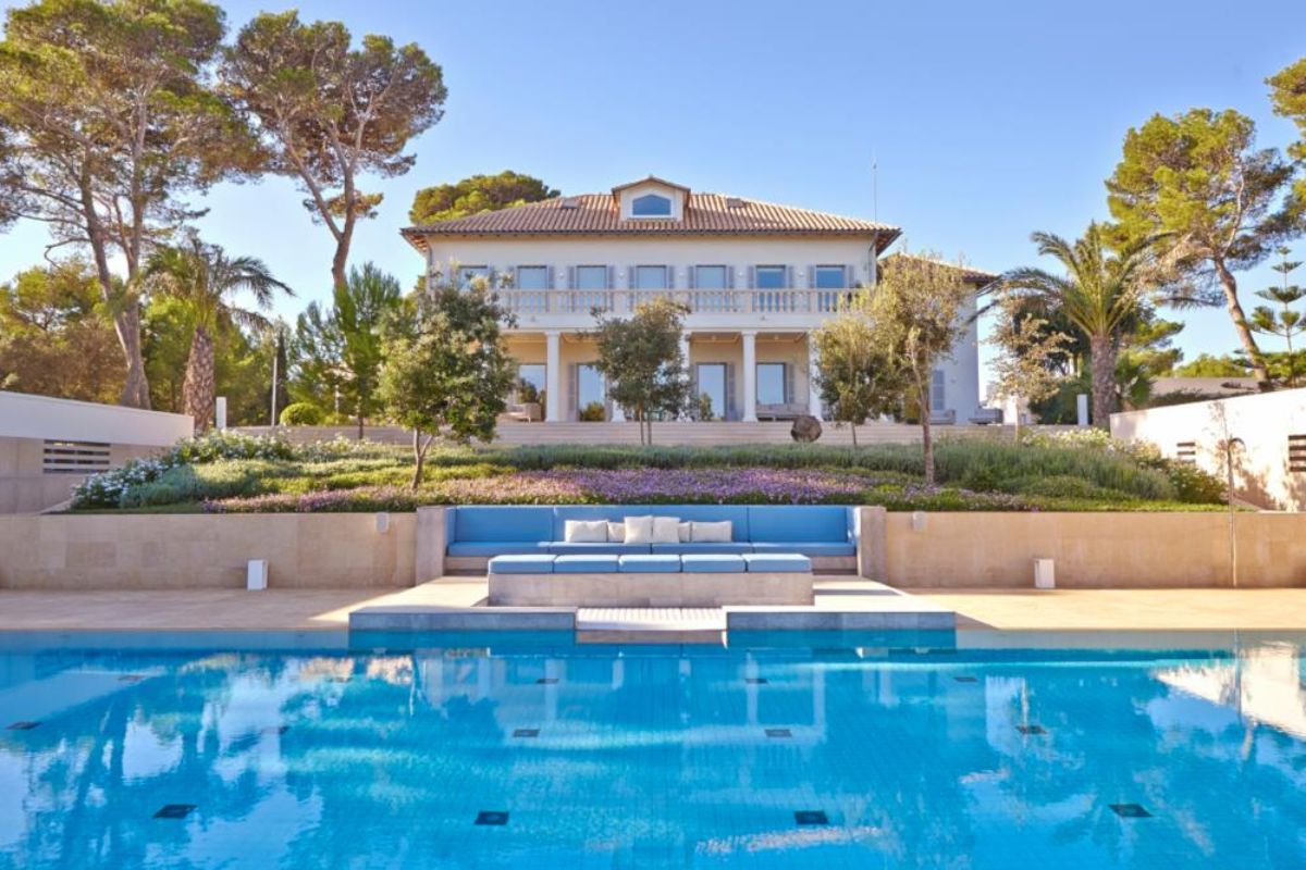 Villa Els Llorers is a large, elegant house with white columns and a tiled roof, set behind a landscaped garden and blue swimming pool, with an outdoor seating area surrounded by tall trees under a clear blue sky.