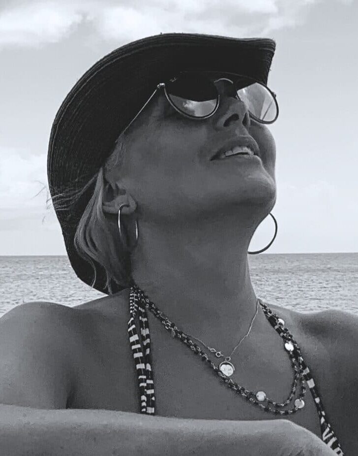 A woman wearing sunglasses, a wide-brimmed hat, and jewellery looks up and smiles by the sea in Barbados. The photo is in black and white.