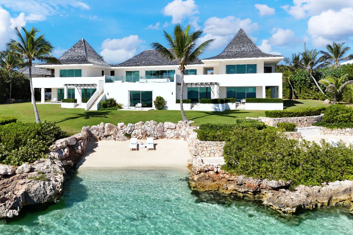 La Bleu, a luxurious white villa with large windows and peaked roofs, sits behind palm trees facing a small private beach and clear turquoise water, surrounded by rocky edges and lush greenery under a bright blue sky.