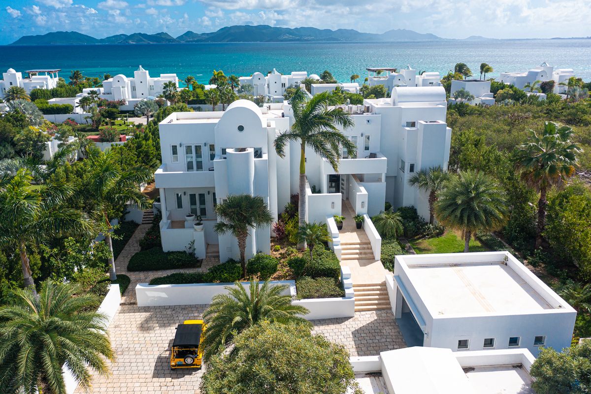 Aerial view of a modern white Shiloh villa surrounded by palm trees, with a yellow vehicle parked outside; the property overlooks turquoise ocean waters and distant mountains under a partly cloudy sky.