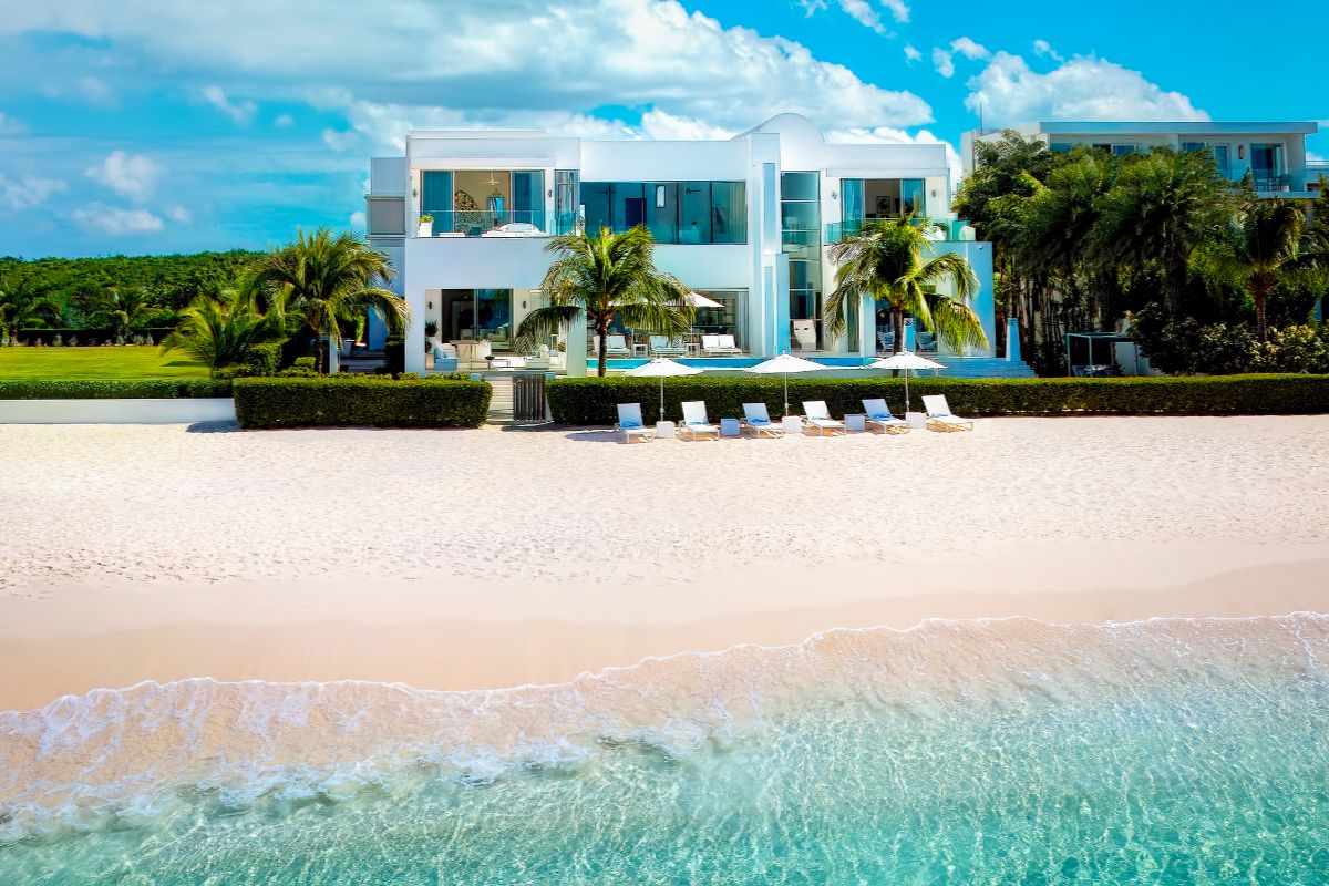 Sandcastle Villa is a modern white beachfront retreat with large windows, surrounded by palm trees, set behind lounge chairs on sandy shores beside clear turquoise water under a bright blue sky with scattered clouds.