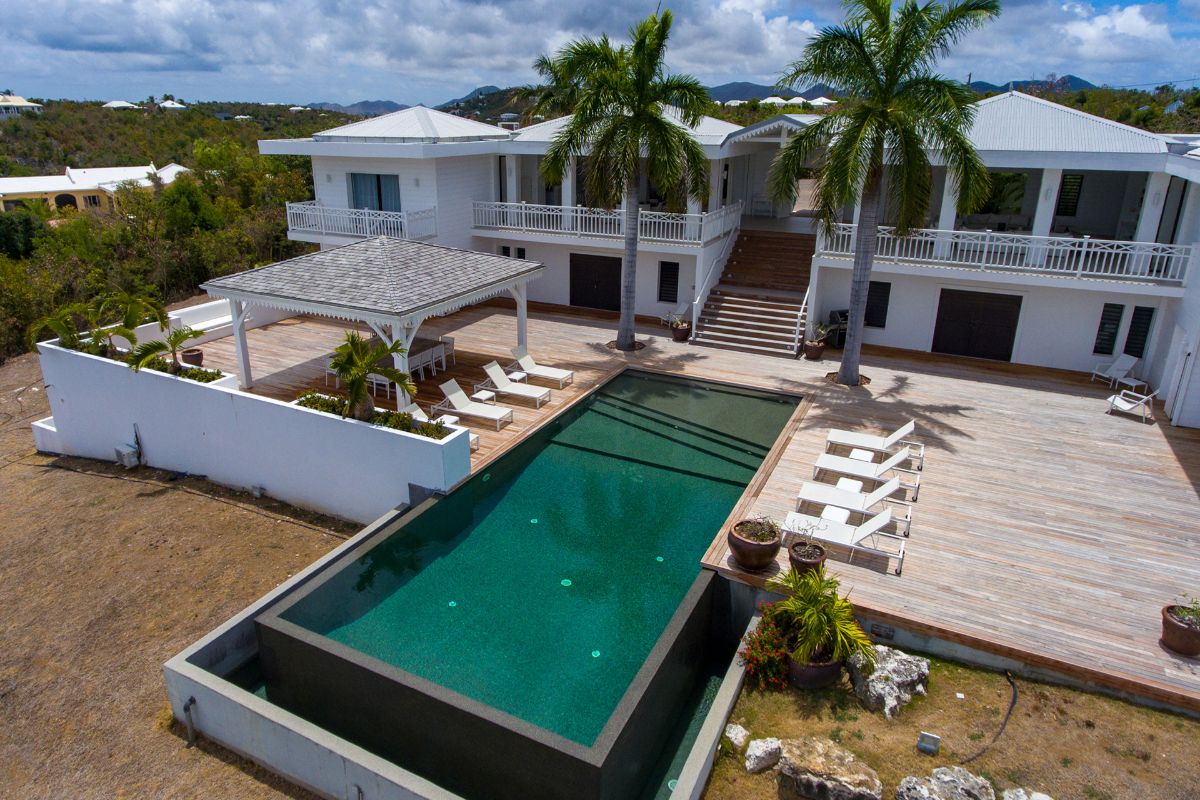A modern white villa, Le Caprice, features large balconies, palm trees, a wooden deck, lounge chairs, and an infinity pool overlooking a scenic landscape under a partly cloudy sky.