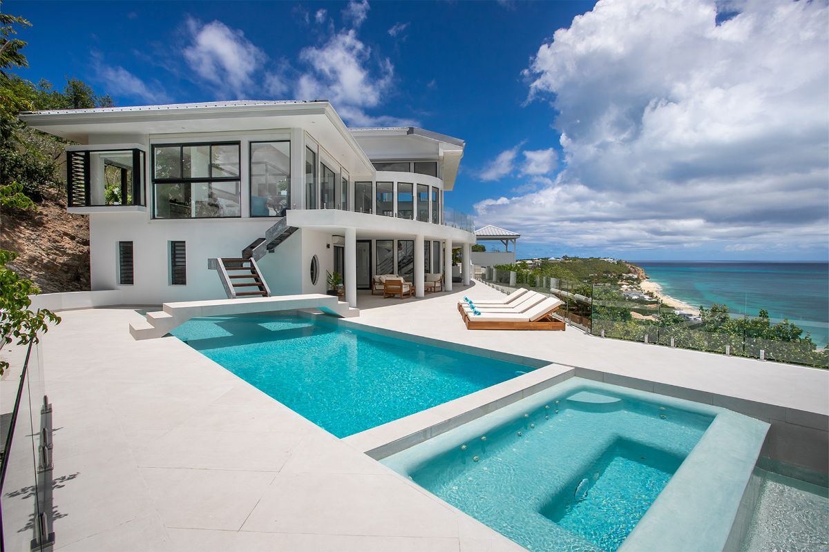 A modern white villa with large glass windows overlooks the ocean and stunning Aqua Rock formations. The patio features a swimming pool, hot tub, two loungers, and clear blue sky with scattered clouds above a scenic coastline.