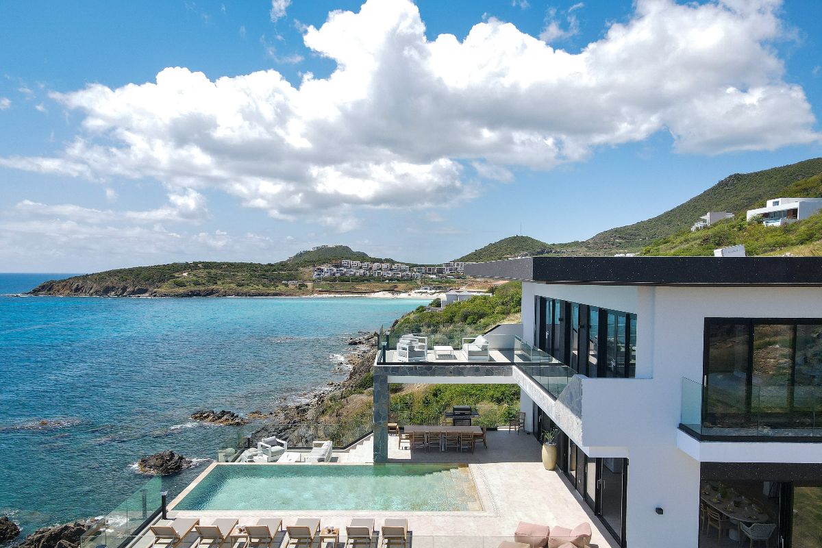 Modern seaside villa with a pool overlooking the Aqua Rock coastline and turquoise ocean, surrounded by green hills under a partly cloudy blue sky. Outdoor lounge chairs line the poolside terrace.