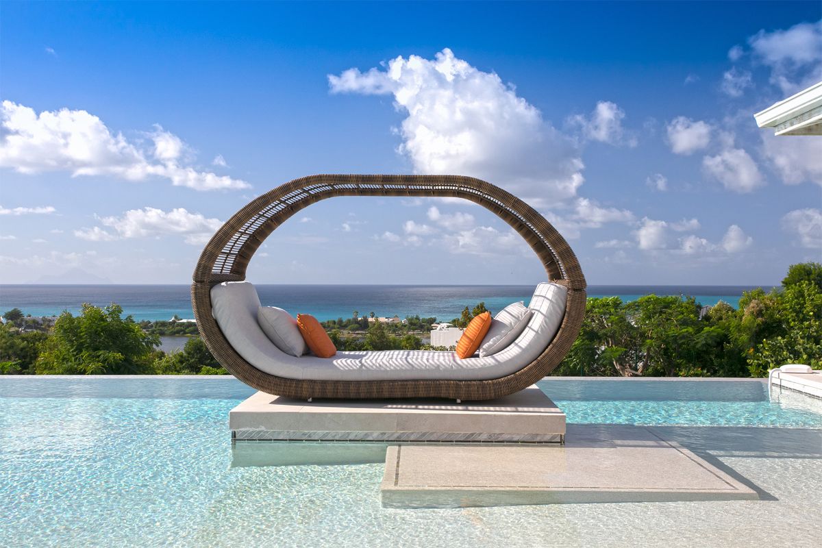A stylish Aqua Vista outdoor daybed with white cushions and orange pillows sits on a platform in a clear infinity pool, overlooking lush greenery and a blue ocean under a bright, partly cloudy sky.