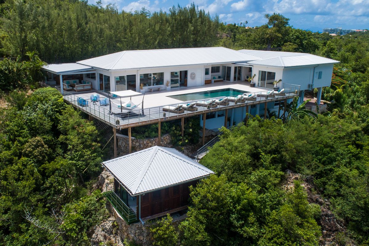 A modern white Avanti villa with a large deck, pool, and lounge area sits elevated among lush green trees, with a smaller building below and distant city views under a partly cloudy sky.