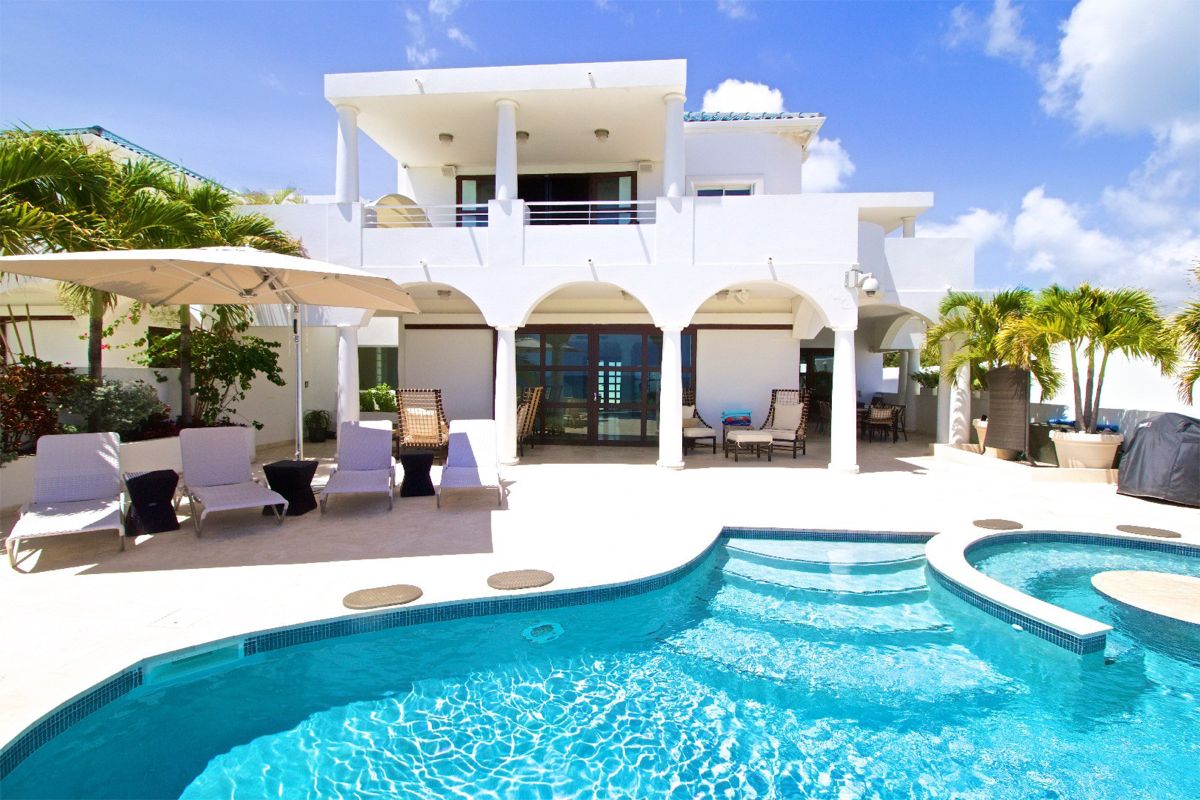 Azur Dream: A modern white two-story villa with balconies, surrounded by palm trees, features a swimming pool, sun loungers, and a large patio umbrella under a bright blue sky.