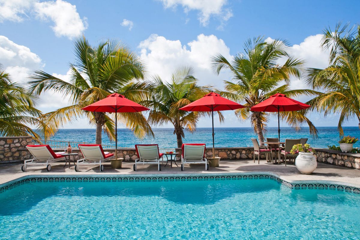 A tropical Bahari poolside scene with lounge chairs and red umbrellas, palm trees swaying, and a stone wall overlooking the ocean beneath a blue sky scattered with clouds.