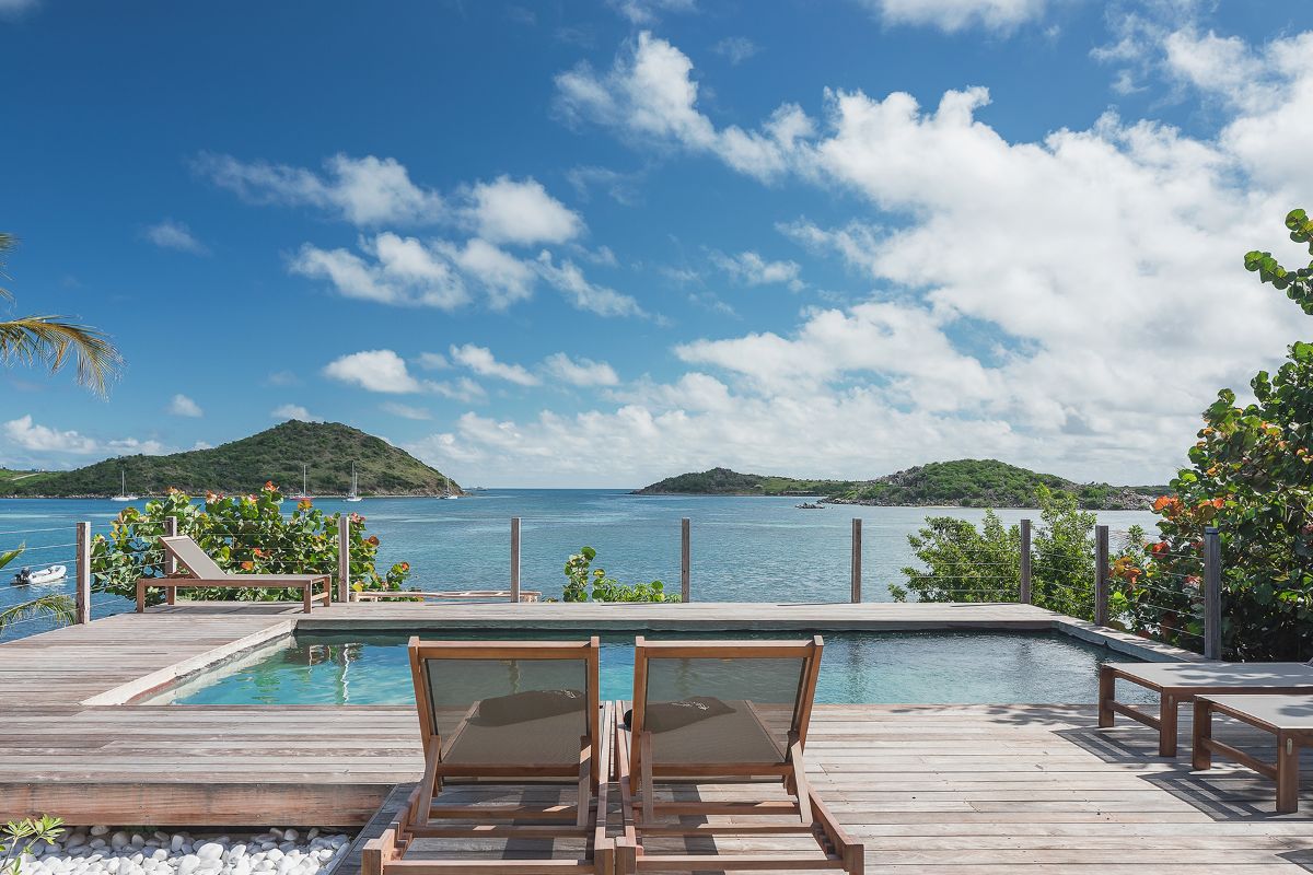 Two wooden lounge chairs sit by a clear pool on a wooden deck at Baie Longue Beach House, overlooking a calm blue sea with green islands in the distance under a bright sky with scattered clouds. Lush greenery surrounds the area.