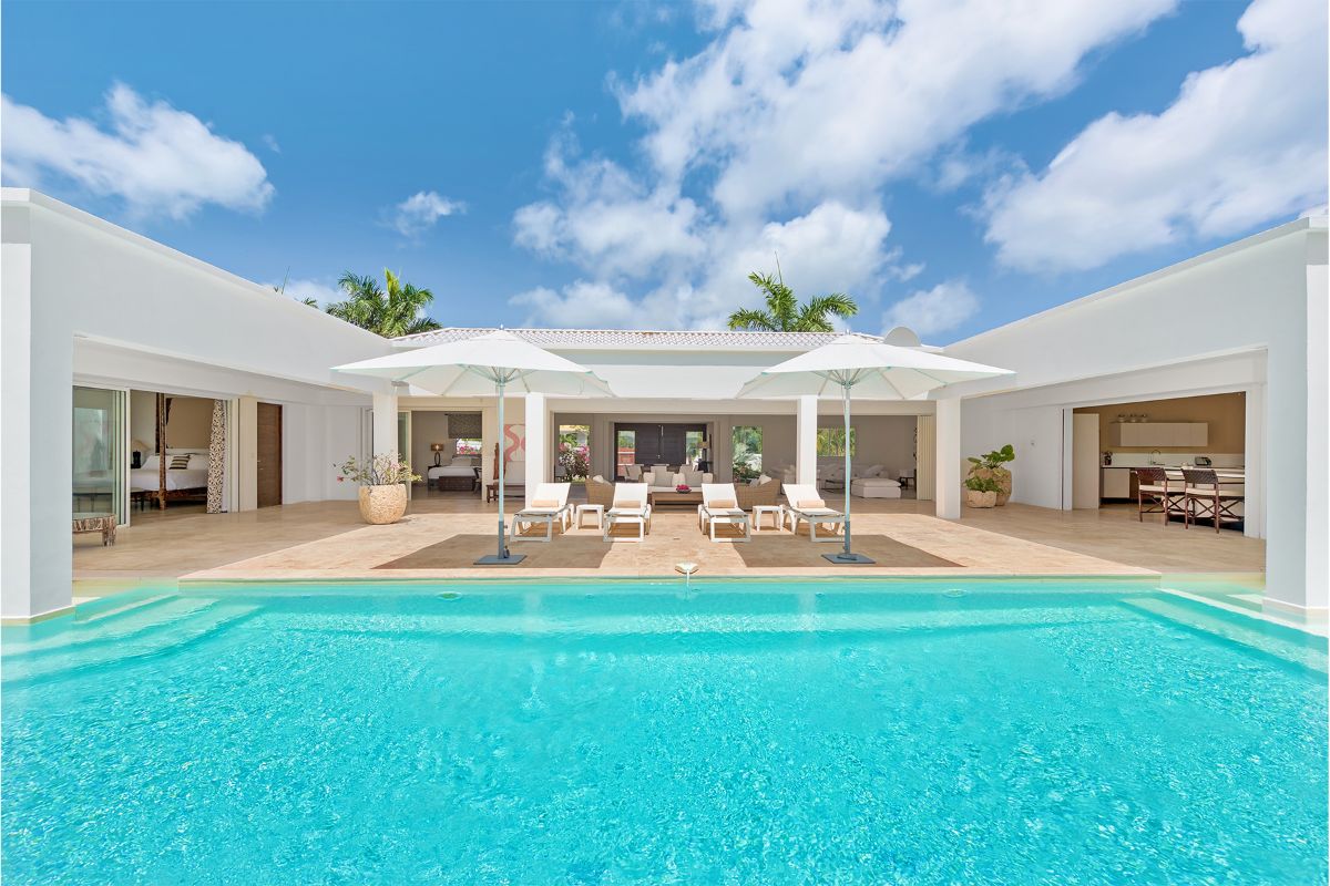A modern luxury villa, Baie Longue Beach House, features an outdoor pool, sun loungers, and white umbrellas with open living spaces and large sliding glass doors under a bright blue sky with scattered clouds.