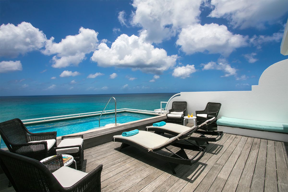 A deck with lounge chairs, a small pool, and wicker furniture overlooks the Blue Beach’s turquoise ocean under a bright blue sky. Towels and drinks on tables enhance the relaxing, luxurious atmosphere.