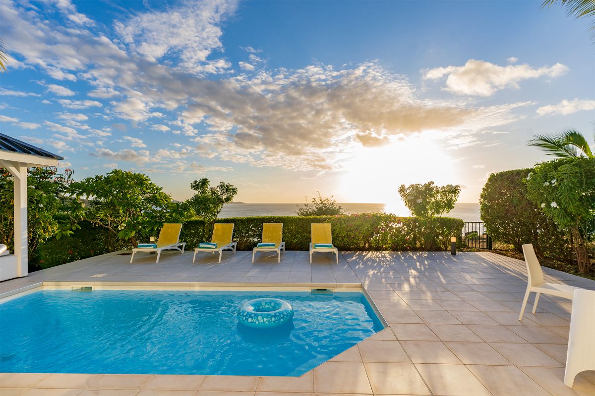 A small pool with a floating ring sits on a tiled patio, surrounded by five yellow lounge chairs. Green shrubs and palm trees border the area, while a blue dove glides above the ocean view at sunset in the background.
