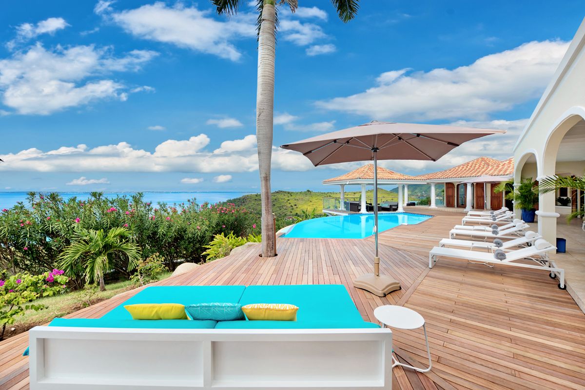 Luxurious outdoor pool area designed for perfect Farniente, with wooden deck, lounge chairs, daybed, and umbrella overlooking lush greenery and blue ocean. White arches and tiled roofs line the villa under a bright, partly cloudy sky.
