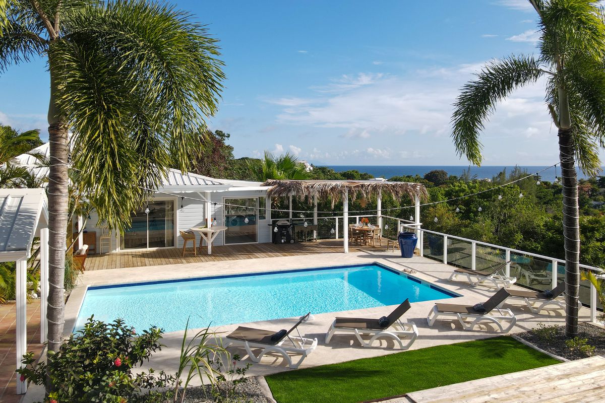 A modern villa at Happy Bay with a rectangular swimming pool, sun loungers, palm trees, and a shaded outdoor dining area, overlooking lush greenery and the ocean under a blue sky with scattered clouds.