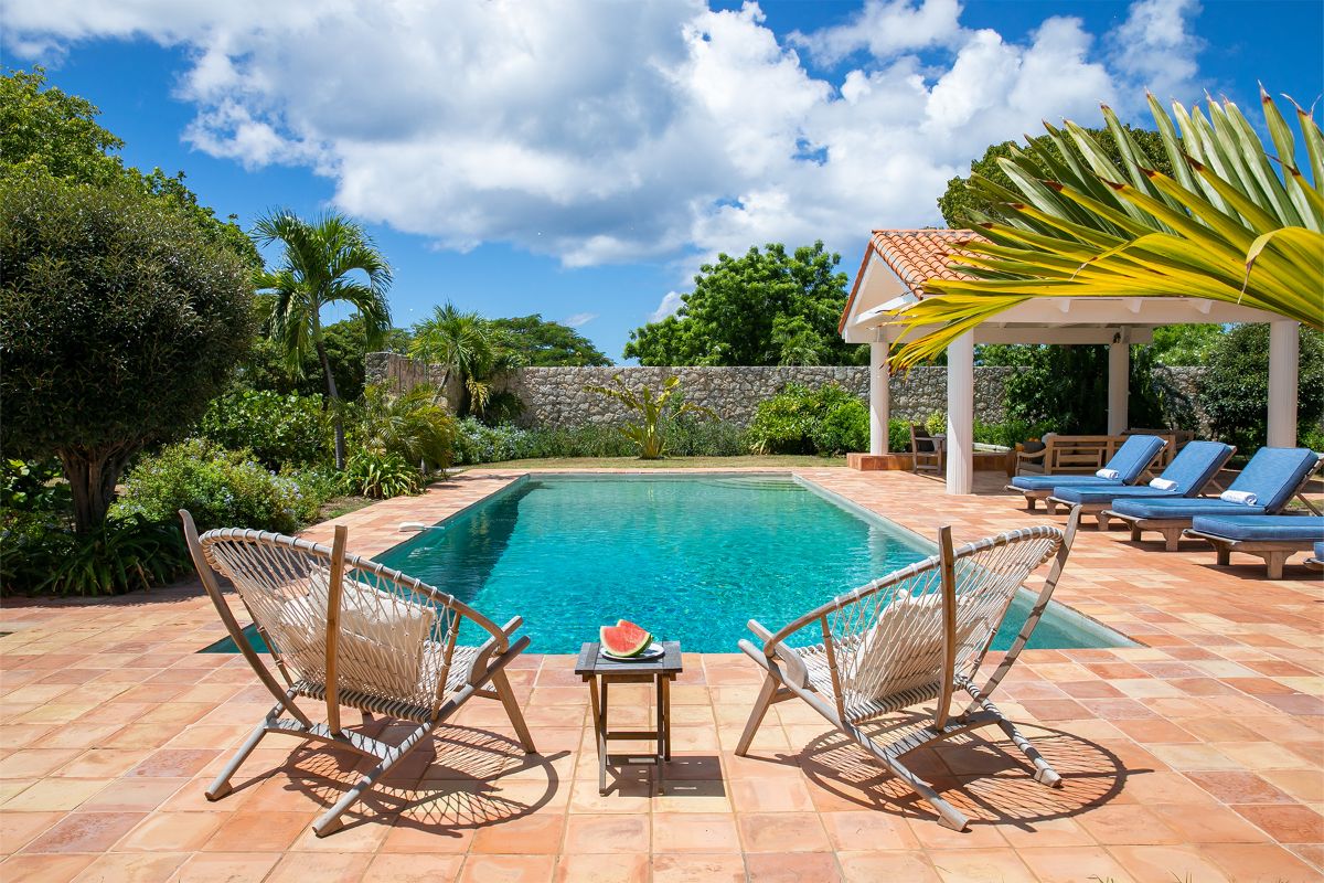 Two wicker chairs and a small table with watermelon sit by a clear swimming pool at Happy Bay, surrounded by lush greenery, lounge chairs, and a covered patio under a bright blue sky with scattered clouds.