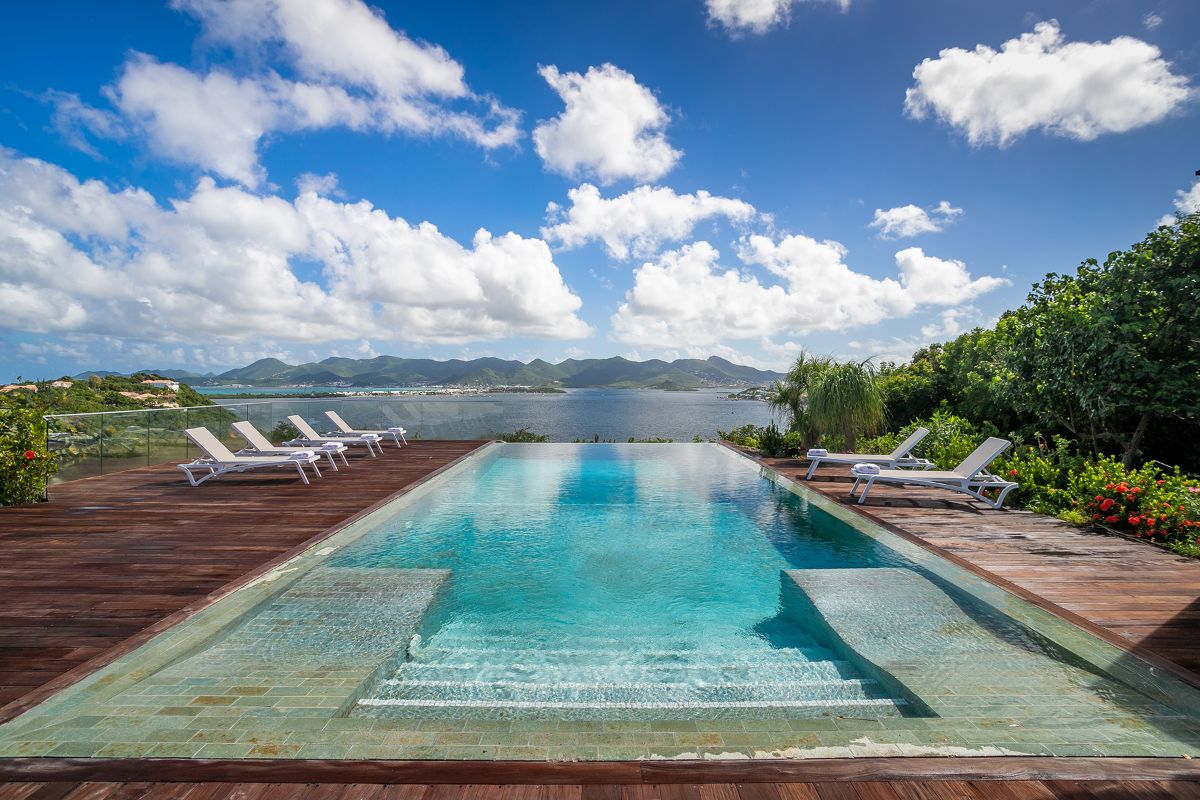 A rectangular infinity pool with clear blue water overlooks Jasper’s scenic lake and distant mountains, surrounded by a wooden deck with white lounge chairs and lush greenery under a partly cloudy sky.