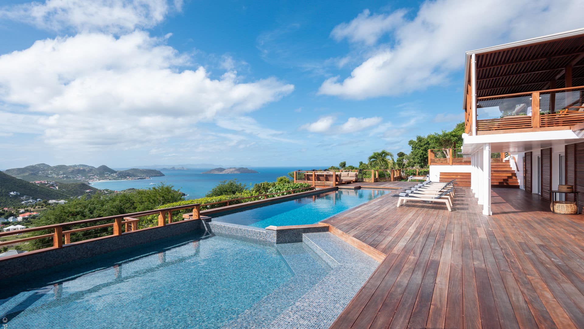 A luxurious villa with a wooden deck, infinity pool, and lounge chairs overlooks a scenic coastline with lush hills and blue ocean under a partly cloudy sky.