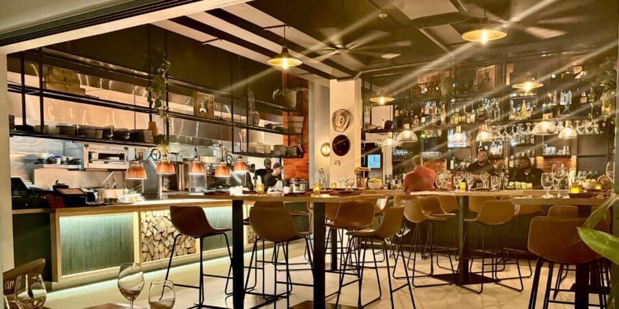 A cosy, modern restaurant with warm lighting, a bar lined with bottles, high tables and chairs, open kitchen area, and decorative plants hanging from the ceiling—just what you’d expect from one of the best restaurants in Turks and Caicos. Some people are seated at the bar.