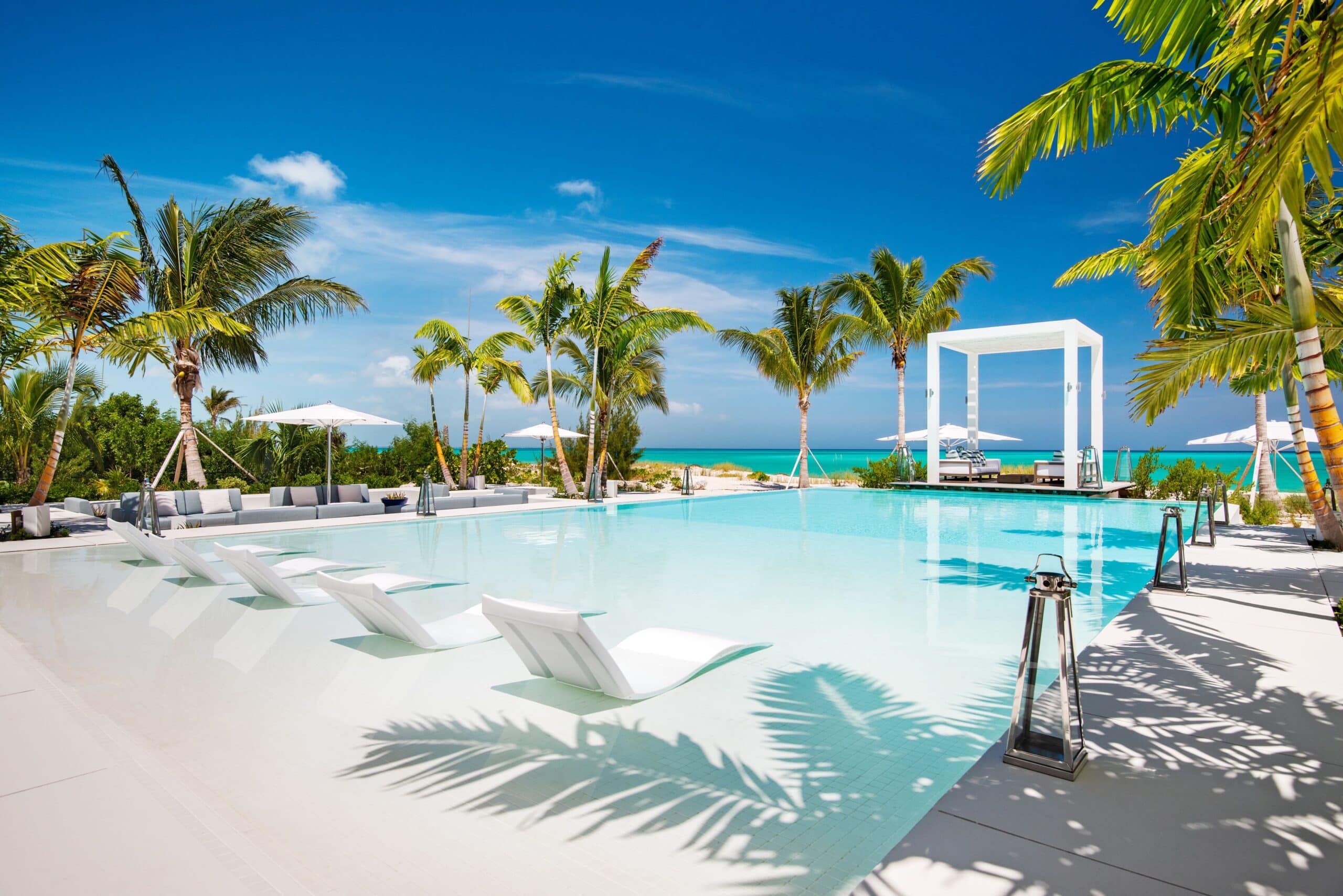 A luxurious outdoor swimming pool surrounded by palm trees, white sun loungers partially submerged in the water, and a white cabana overlooking a sandy beach and turquoise sea under a clear blue sky.