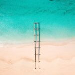 Aerial view of a wooden pier extending from a sandy beach near luxury villas Turks and Caicos, stretching into clear turquoise water with gentle waves and dark patches visible beneath the surface.