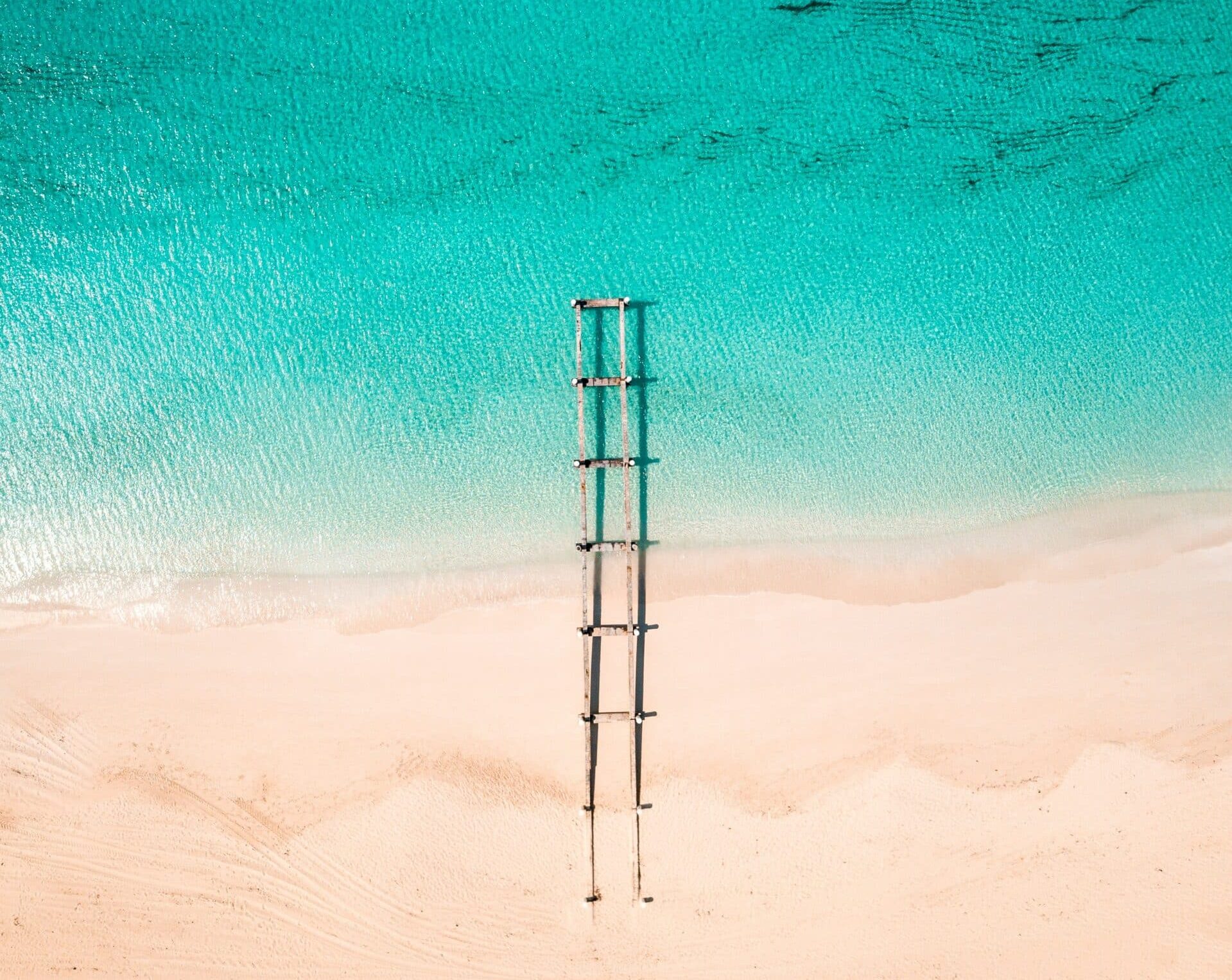 Aerial view of a wooden pier extending from a sandy beach near luxury villas Turks and Caicos, stretching into clear turquoise water with gentle waves and dark patches visible beneath the surface.