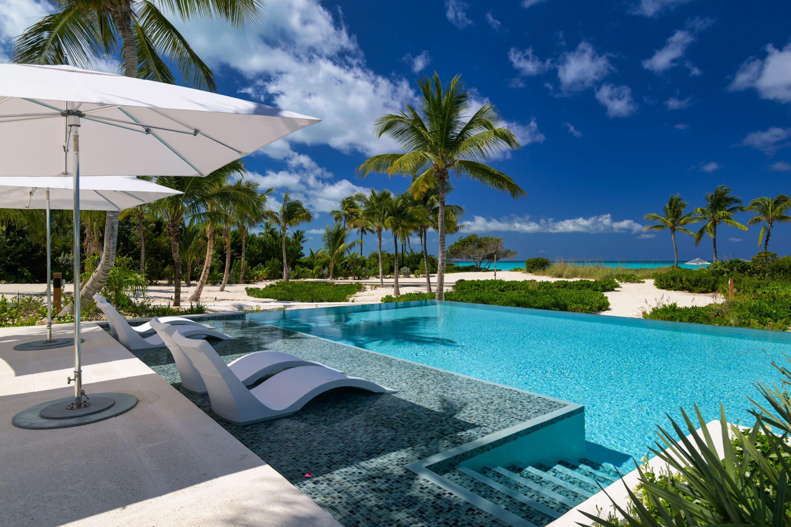 A luxurious infinity pool with white loungers overlooks a sandy beach lined with palm trees under a bright blue sky—perfect for your luxury beachfront villa hire in Turks and Caicos. Large white parasols provide shade poolside.