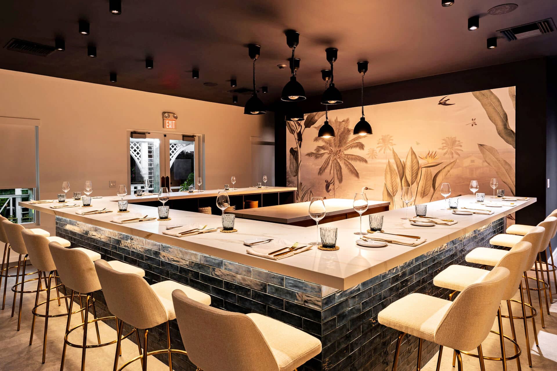 Modern U-shaped dining bar with cushioned bar stools, set with plates, glasses, and cutlery, under black pendant lights. A mural with plants decorates the back wall—stylish touches found in the best restaurants Turks and Caicos has to offer.