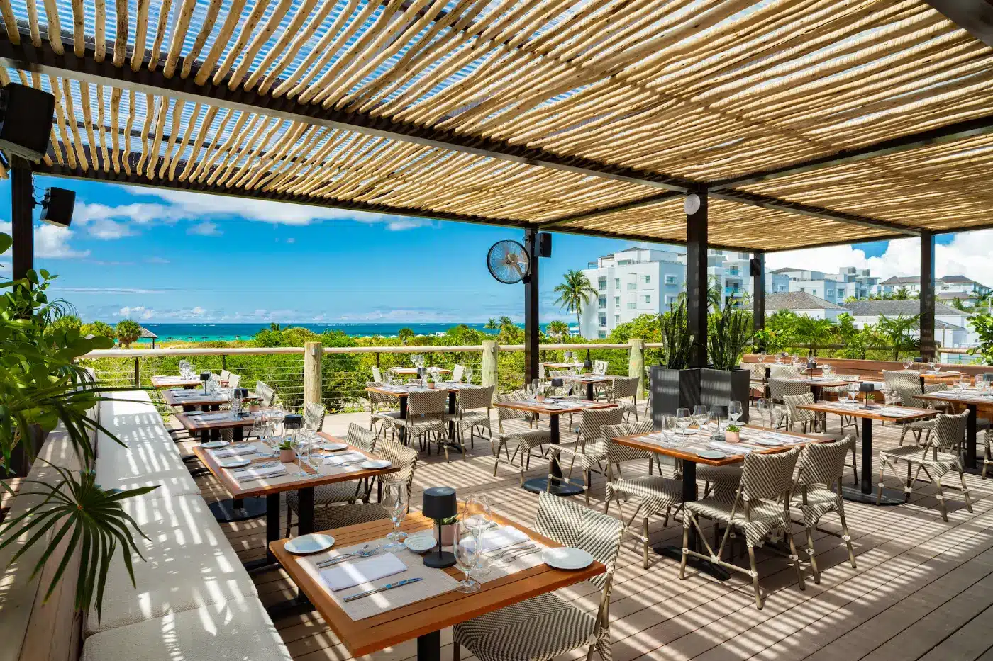 Outdoor restaurant terrace with wooden pergola, neatly set tables and chairs, lush plants, and a view of the sea—one of the best restaurants Turks and Caicos offers, surrounded by white buildings under a blue sky with scattered clouds.