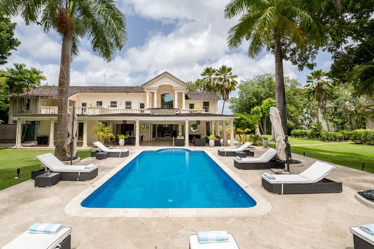 Large luxury Sandy Lane Villa with Cottage featuring a back garden pool, surrounded by palm trees and sun loungers on a sunny day. The cream-coloured house boasts two storeys, balconies, and well-kept lawns, creating a relaxing tropical setting.
