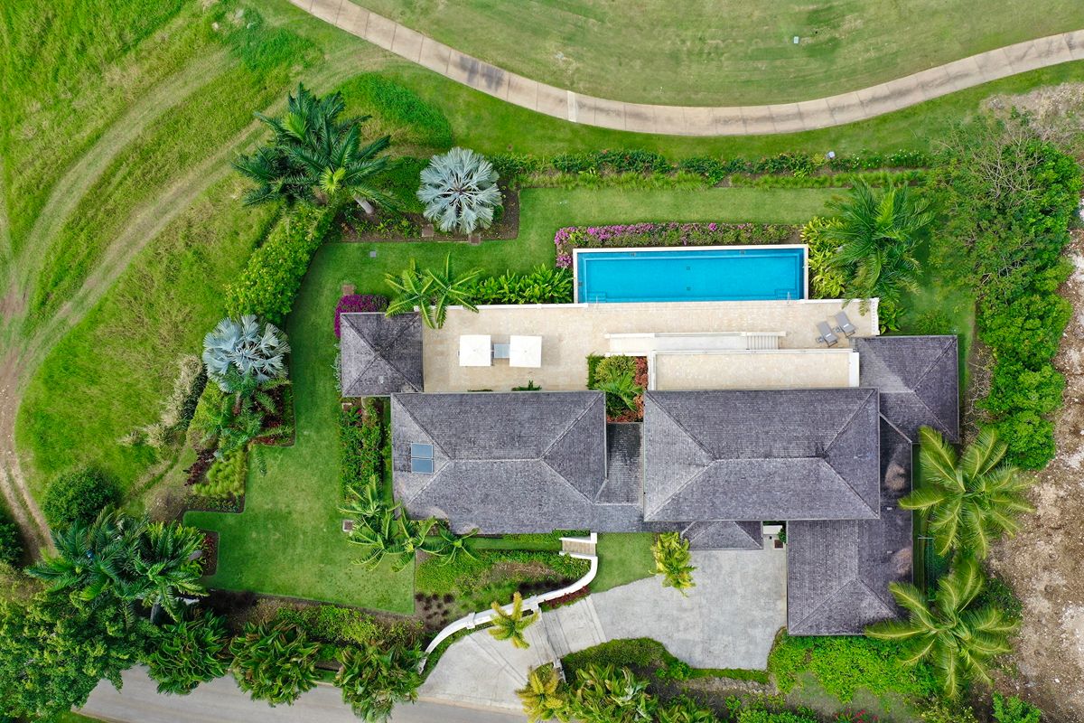 Aerial view of a modern house with a grey roof, a rectangular swimming pool, lush green garden, trees, and surrounding paths—an ideal setting for an Apes Hill Golf Villa Rental.