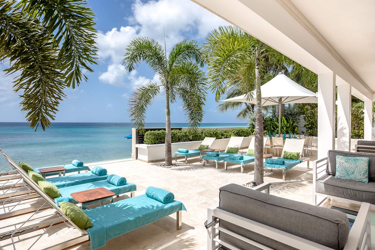 Lounge chairs with turquoise towels and cushions sit on a sunny patio by the sea, surrounded by palm trees and white parasols, with clear blue sky above—an idyllic spot where you might even catch a glimpse of a playful dolphin leaping in the distance.