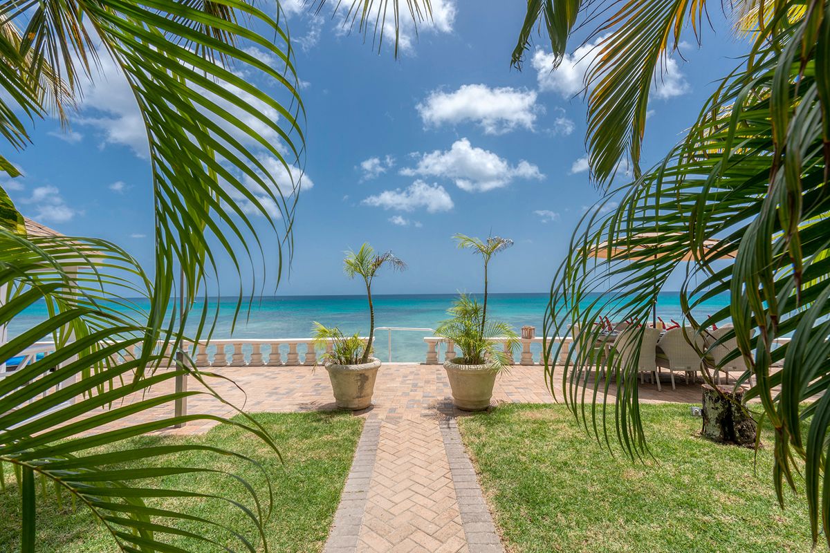 A tropical beach scene at Ebb Tide features a paved path leading to the sea, flanked by palm trees and potted plants. Blue sky, white clouds, and loungers are visible in the background.