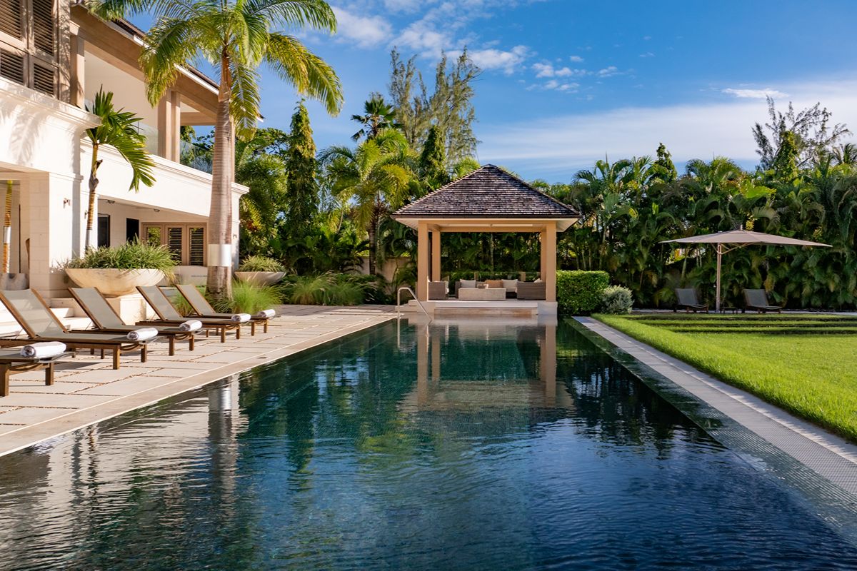 Modern back garden with a rectangular pool, loungers, palm trees, a grassy lawn, and a gazebo reminiscent of the serene style found at Godings Beach House. The sky is clear and sunny, creating a relaxing atmosphere.