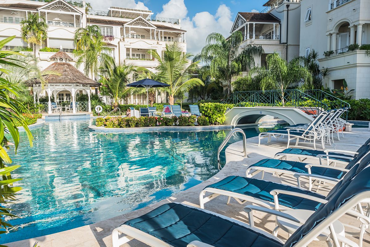 Resort pool area with blue sun loungers, a footbridge over the water, lush palm trees, and elegant white buildings in the background under a blue sky with clouds.
