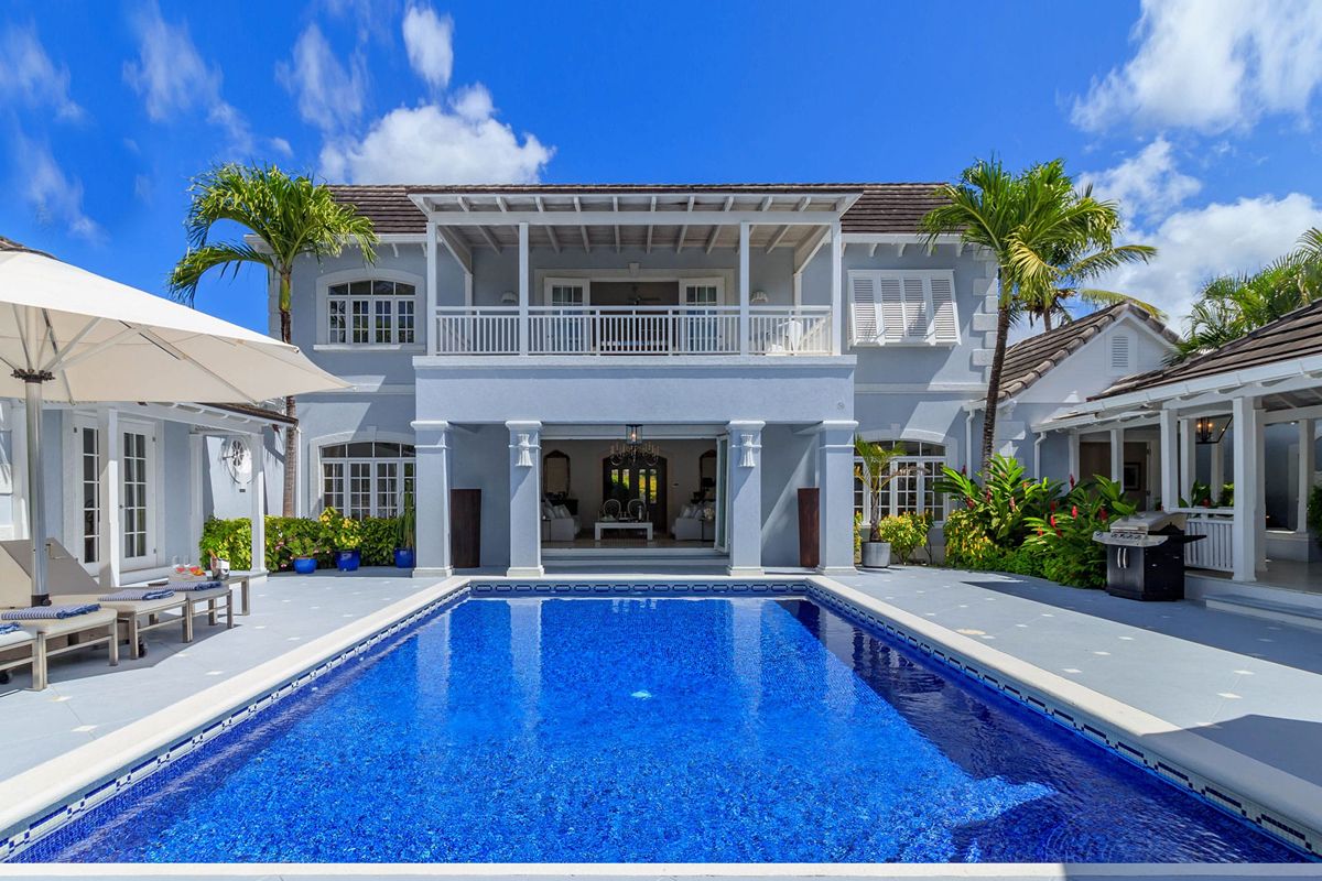 Luxury two-storey house with a large blue swimming pool, loungers, parasols, and lush palm trees swaying gently in the trade winds under a bright blue sky with scattered clouds.
