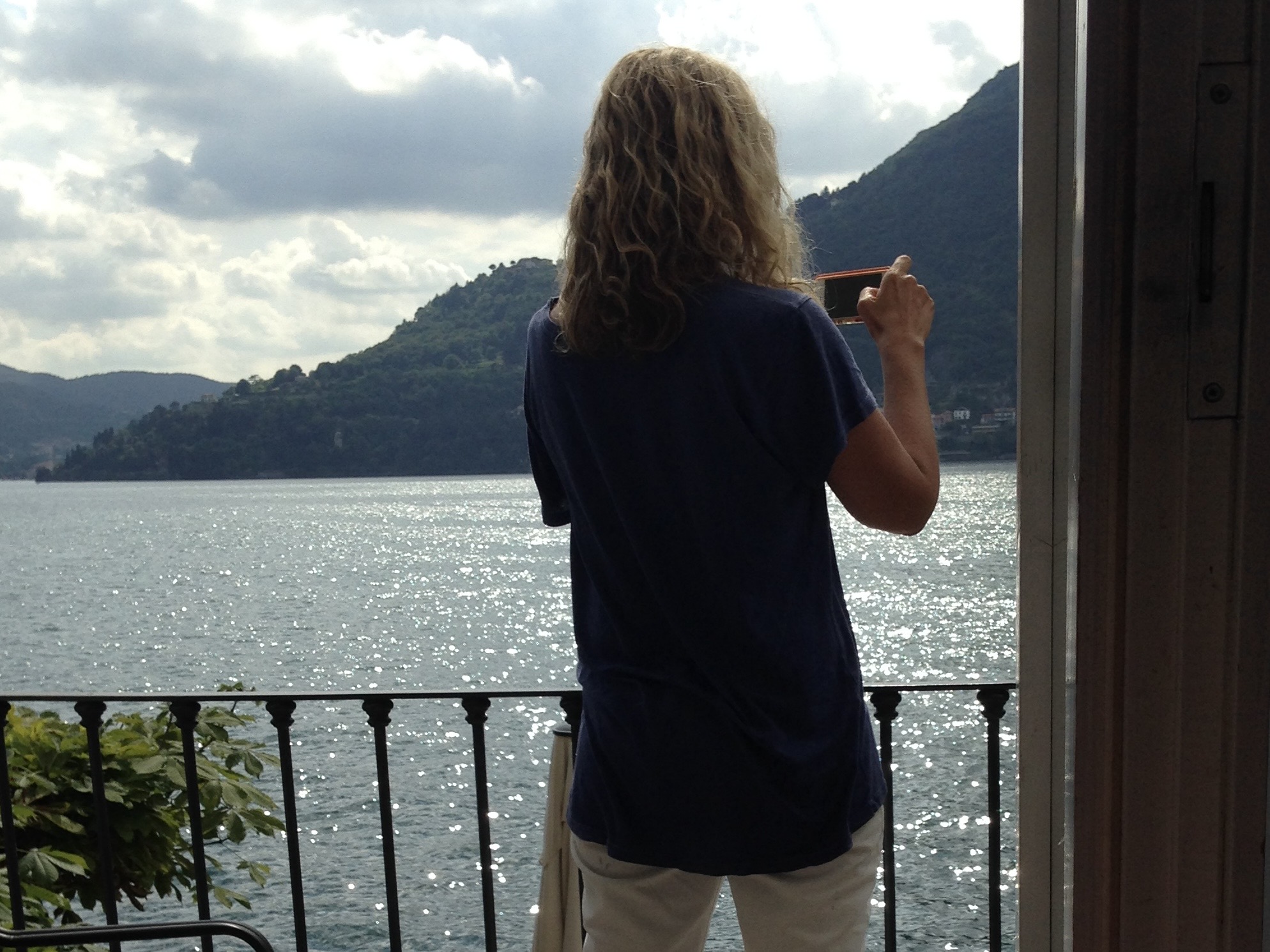 A person with wavy hair stands on the balcony of one of the luxury villa rentals, overlooking a sparkling lake with hills in the background, taking a photo with a mobile phone.