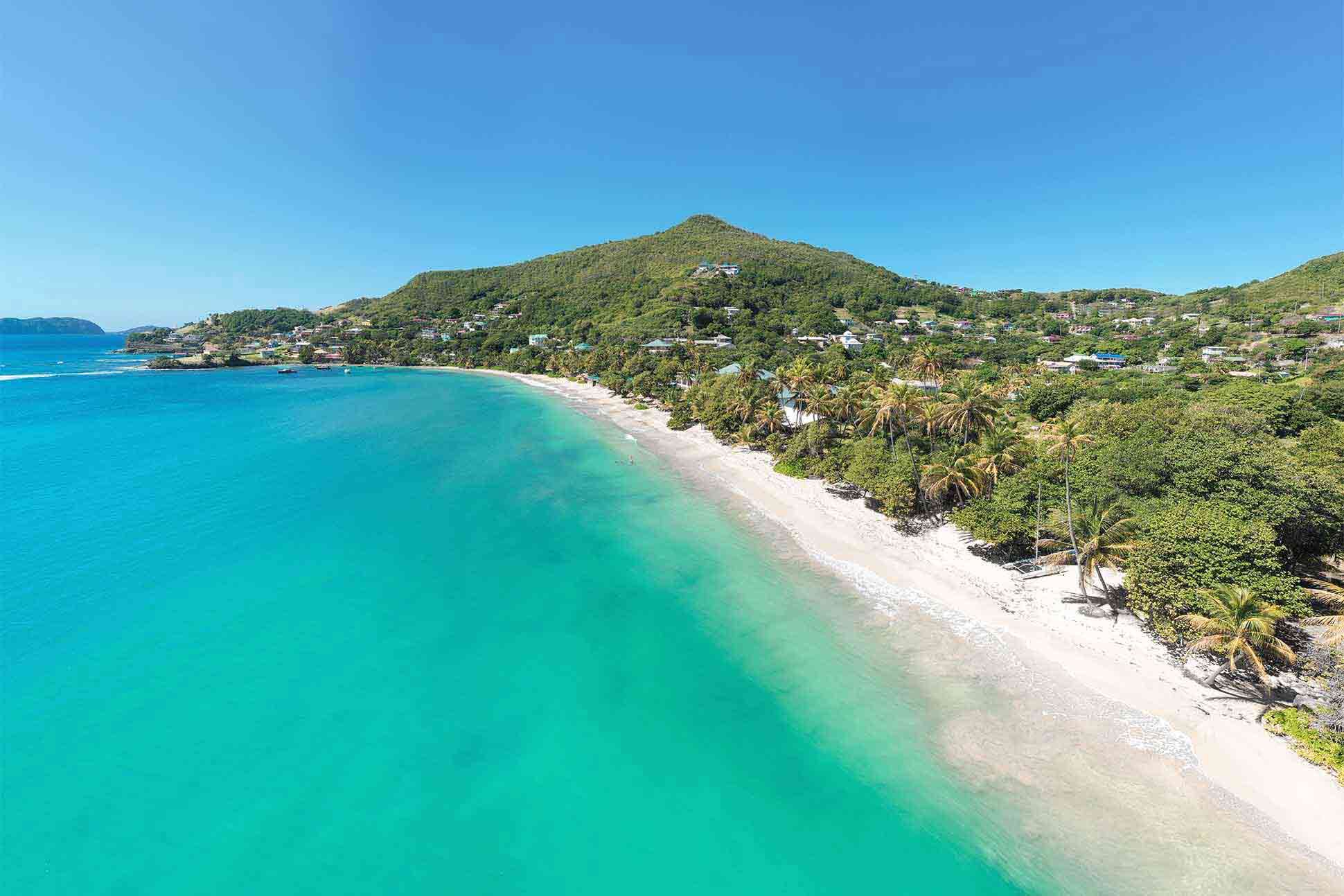 Aerial view of a tropical beach with turquoise water, white sand, palm trees, and lush green hills under a clear blue sky—a scene straight from the pages of a Bequia travel guidebook.