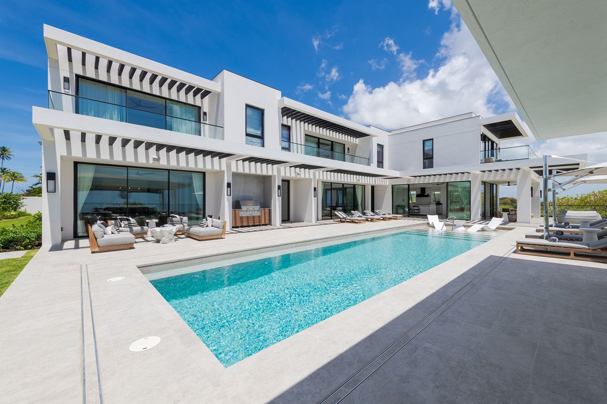 Modern two-storey white villa with large windows, a spacious outdoor patio, lounge seating, and a rectangular swimming pool under a sunny blue sky with scattered clouds.