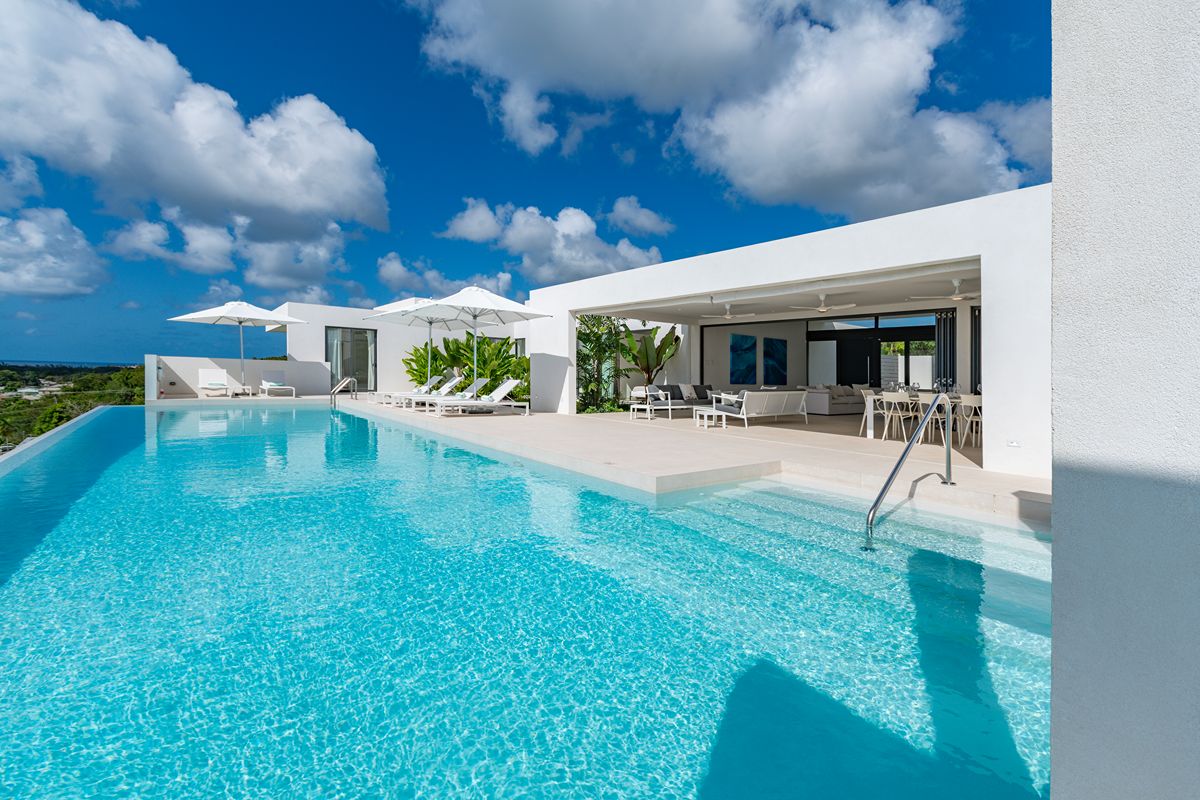 Modern white villa, reminiscent of Villa Palmier Barbados, with a large infinity pool, sun loungers, and umbrellas under a blue sky with fluffy clouds, surrounded by lush green plants and an open lounge area.